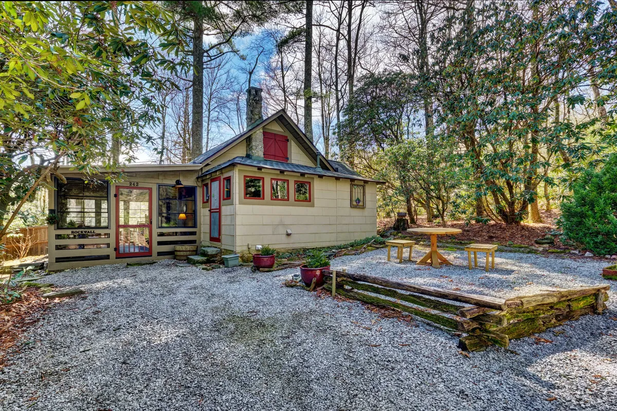 The image shows a quaint and charming cottage nestled in lush greenery, with a gravel pathway leading to a cozy wooden seating area. The sun sets in the background, casting a warm glow over the patio, inviting guests to unwind outdoors or stargaze by the fire pit. The atmosphere is serene and private, perfect for a romantic getaway.