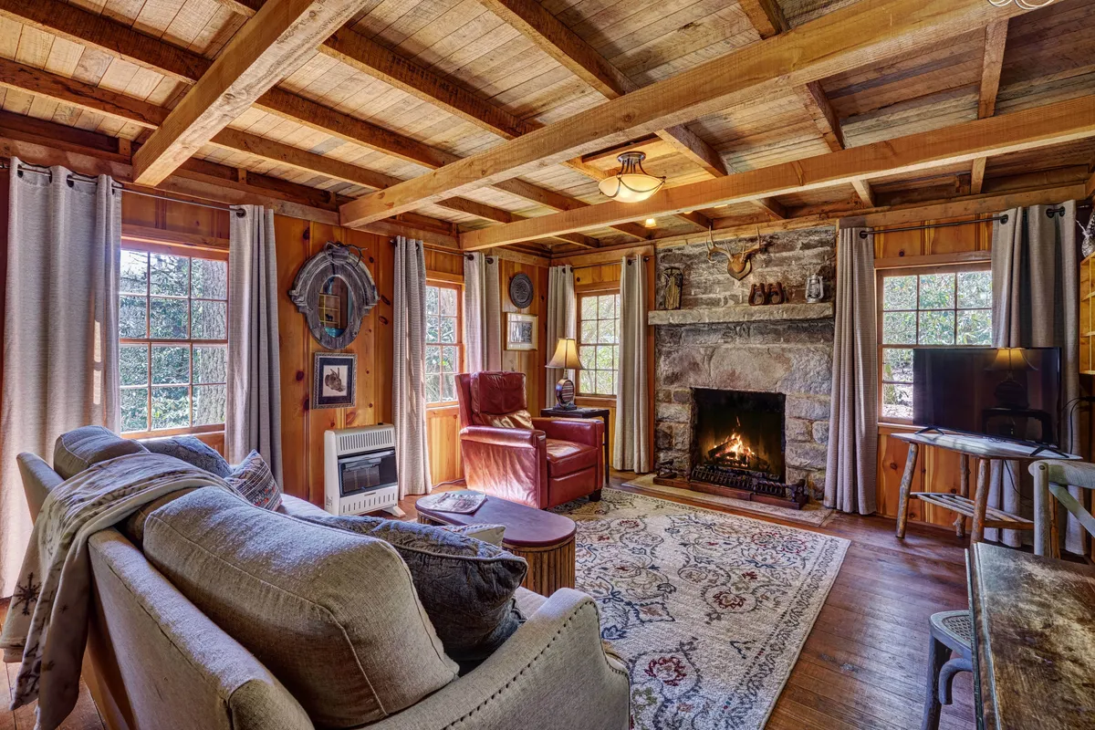 This image presents the heart of the living room, featuring a prominent stone fireplace as the focal point. A comfortable sofa and armchair are arranged around a patterned rug, framed by large windows that invite natural light. The space exudes a welcoming atmosphere, ideal for relaxing, socializing, or enjoying the warmth of the fire. Whimsical decor and antiques subtly accent the shelves and walls, reinforcing the cabin's historic character.