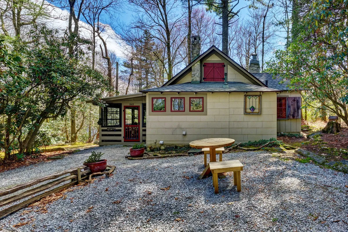 A serene exterior shot of Rockwall Cabin nestled among mature trees, featuring a charming cottage with a round wooden table on the gravel patio. The setting is peaceful and private, ideal for intimate outdoor dining or enjoying morning coffee while surrounded by nature.