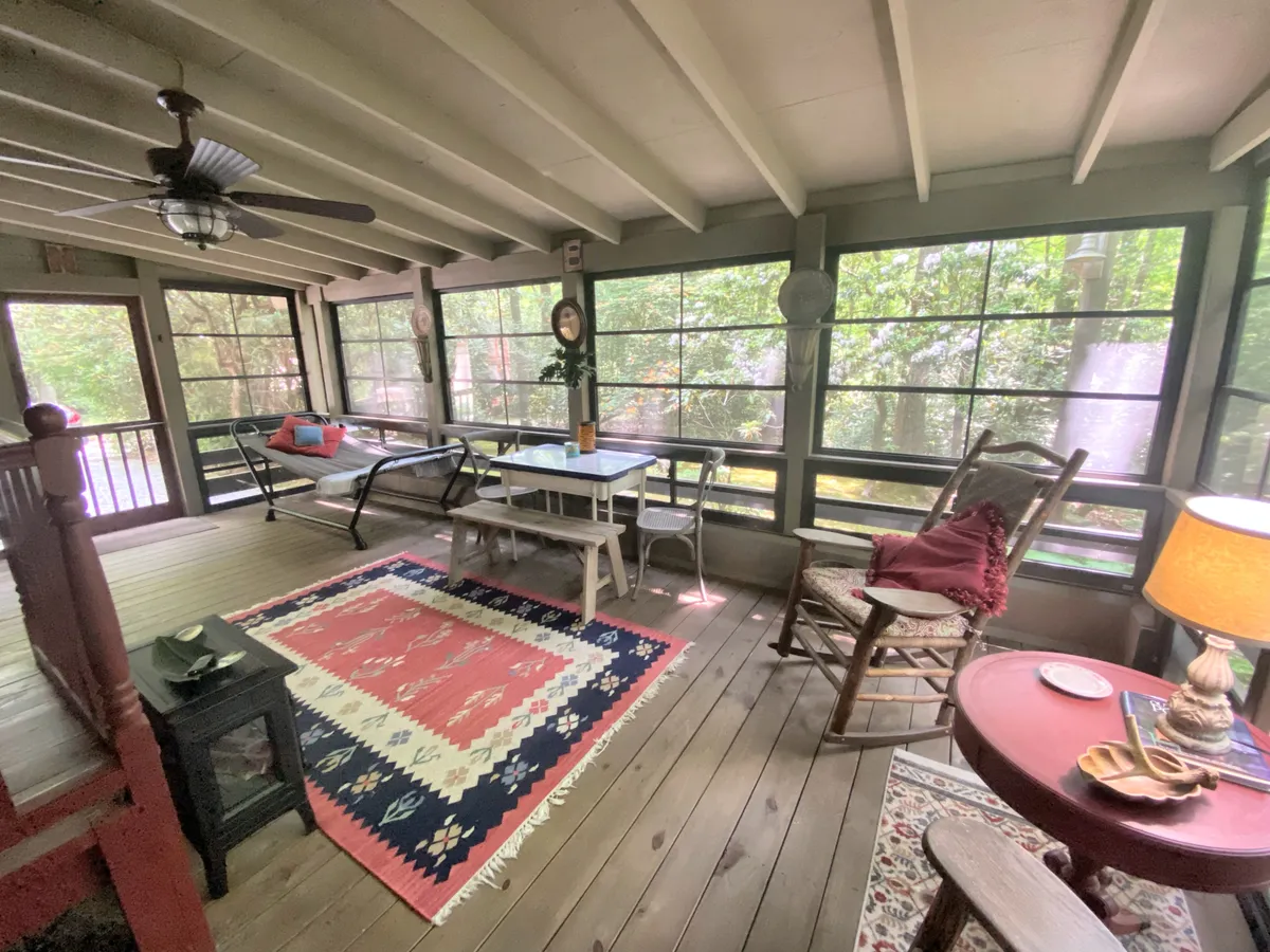 A serene interior shot showcasing large windows framing lush outdoor views, exposed wooden ceiling beams, a colorful area rug, a rocking chair, and a small table with a lamp. The soft lighting and natural materials create a tranquil, welcoming environment ideal for reading or unwinding.