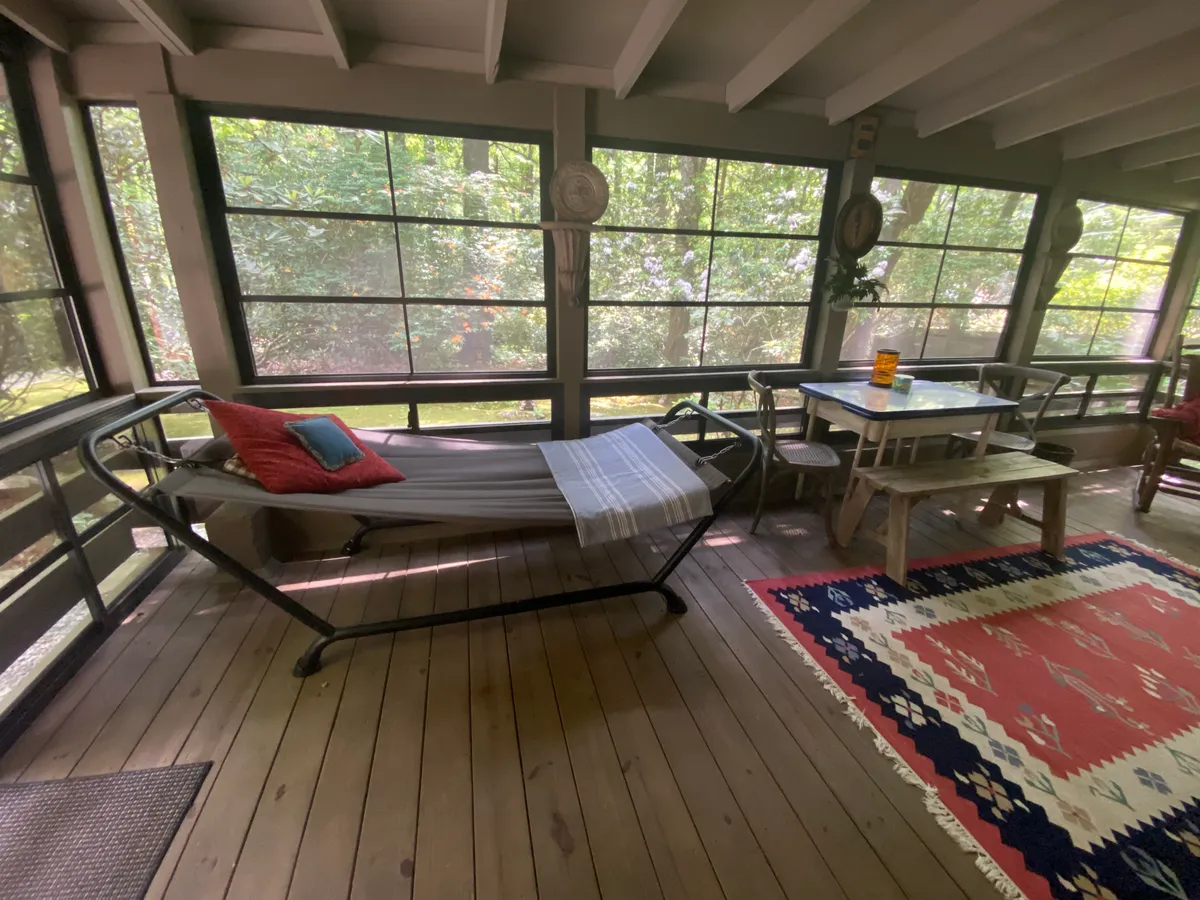 This image features a comfortable hammock strung across a light-filled space, a dining table, and a rug. The room is surrounded by large windows that allow for expansive views of the outdoors, and the airy ambiance suggests an inviting transition between indoor comfort and outdoor scenery.