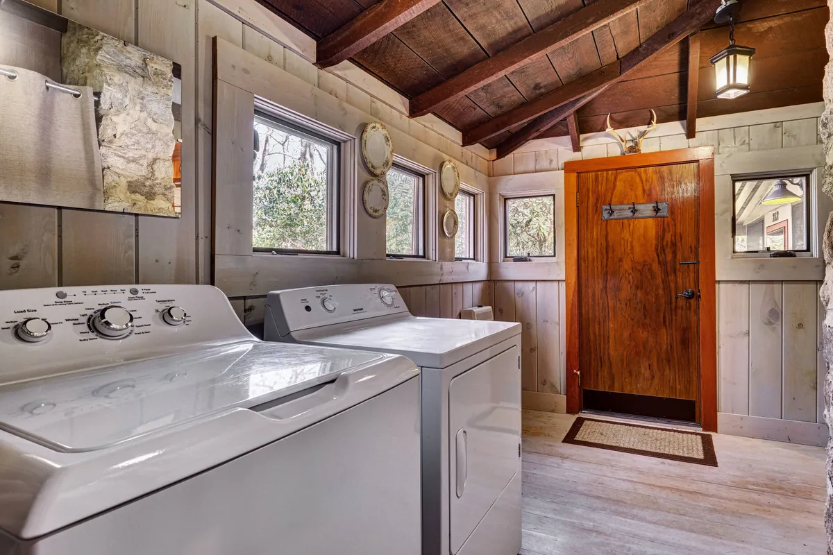 A charming laundry room featuring a vintage-style wooden door, sunlight streaming through the windows, decorative plates adorning the walls, and classic appliances like a washing machine and dryer. The mix of old and new gives the space a nostalgic yet practical feel.