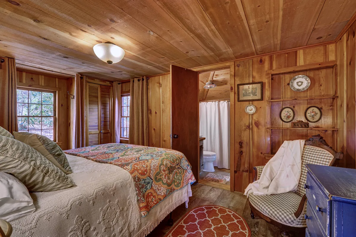 A cozy bedroom with a bed dressed in a colorful quilt, surrounded by rustic wooden paneling, vintage furniture, and whimsical decorative plates on a shelf. Natural light streams in from a window with soft curtains, creating a warm, inviting atmosphere that blends comfort and character.