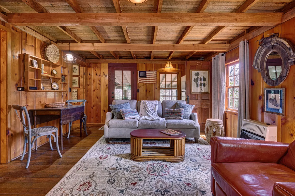A broader perspective of the living area, highlighting a plush sofa, a rustic coffee table, exposed wooden beams, and thoughtfully curated artwork and decor. Shelves display both vintage and playful decorative pieces, continuing the cabin's whimsical historic theme. The arrangement invites relaxation and fosters a sense of creative inspiration.