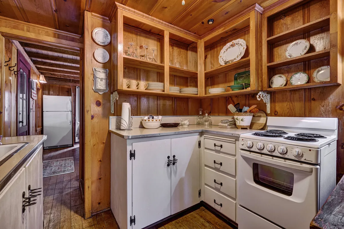 This image showcases the heart of the kitchen, featuring warm wooden cabinetry, a central stove, and open shelving adorned with dishes and glassware. The space radiates a welcoming ambiance, blending modern functionality with vintage character.