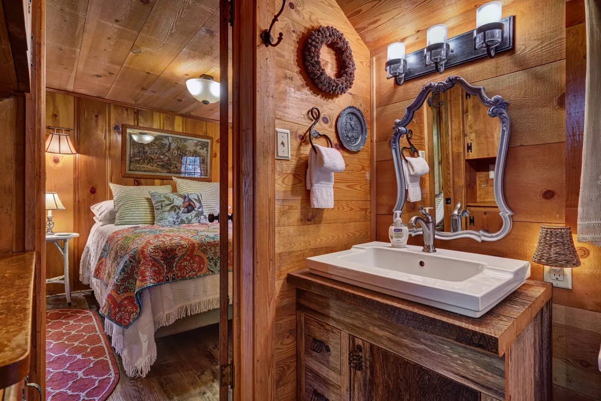 A space blending modern and vintage touches, this image captures a vintage-style mirror above a sink with a modern faucet. The room features wooden paneling, colorful bedspread glimpses, and artwork, reflecting a bath area with both contemporary comfort and antique charm.