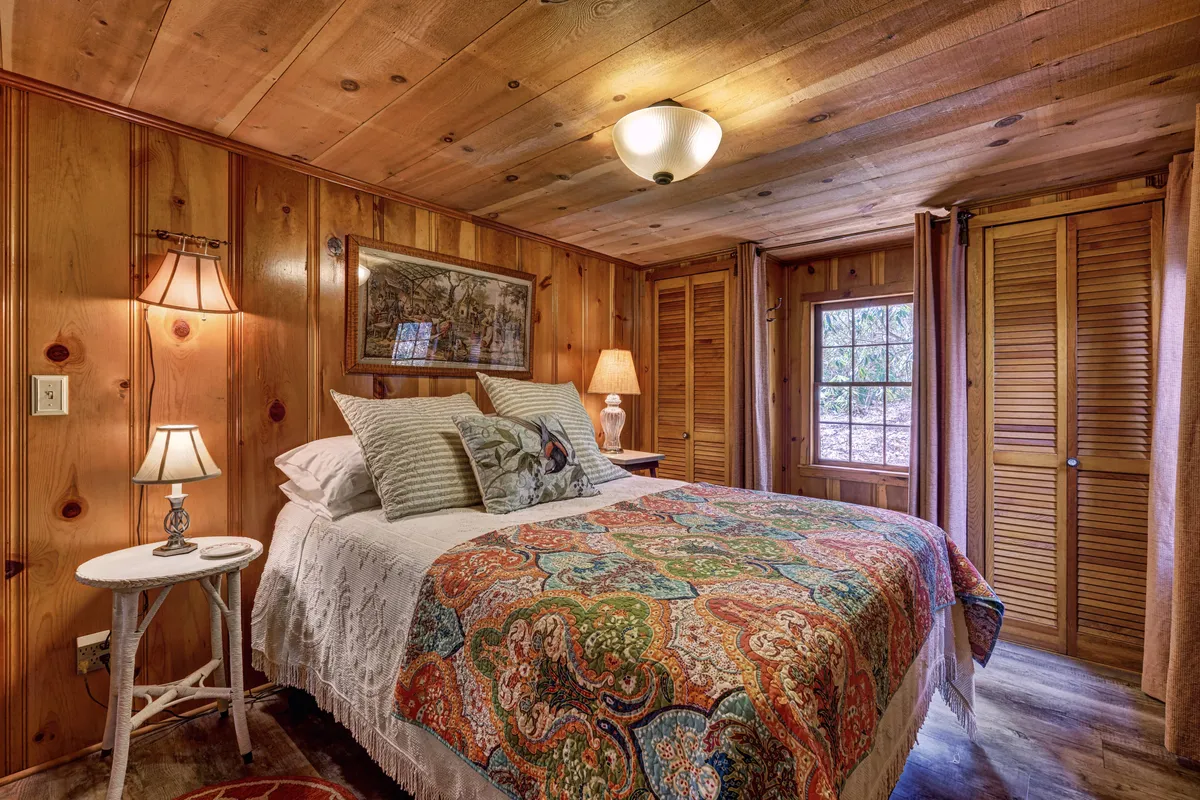 Another view of the bedroom, featuring a bed with a colorful quilt, a bedside table lamp, framed artwork on the wall, and wooden closet doors. A window offers a glimpse of the outdoors, reinforcing the connection to the surrounding forest and the tranquil ambiance of the retreat.