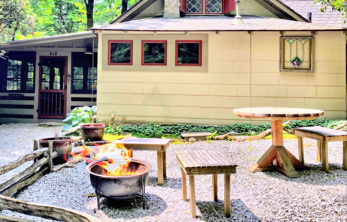 An inviting exterior view of the cabin’s outdoor fire pit area, bordered by lush greenery and accented by a stained glass window. Wooden tables surround the fire pit, suggesting an ideal setting for gathering, sharing stories, or roasting marshmallows under the stars.
