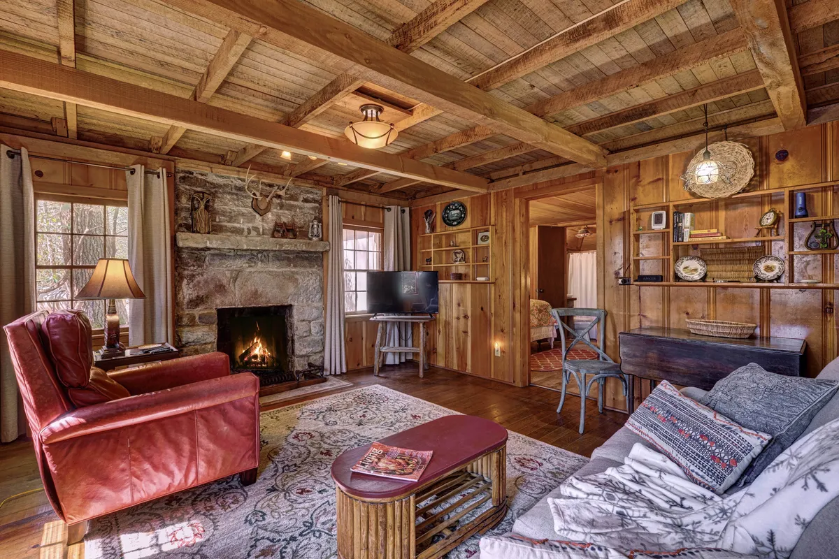 This view focuses on the restored fireplace, accented by a bold red armchair, a textured rug, and shelves filled with books and antiques. The wooden floor gleams with character, and eclectic decor items lend personality and a sense of history to the space. The overall ambiance is both intimate and inviting, emphasizing the cabin’s unique charm.