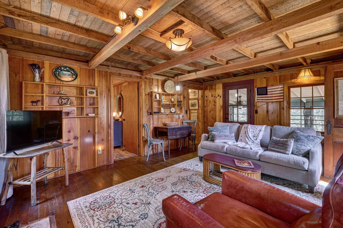 A cozy living area featuring a comfortable sofa, wooden shelves lined with books and decorative items, a rustic dining table, and a plush rug anchoring the space. The ambiance is warm and inviting, with natural light filtering through windows and a sense of quietude that encourages relaxation and introspection.